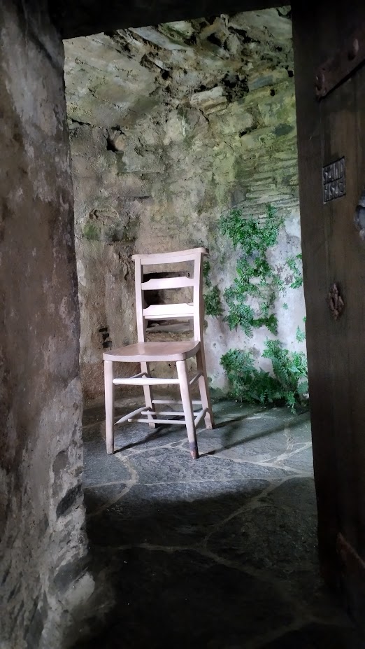 Photographie d'une chaise prise lors d'une visite de l'abbaye de Iona en Écosse. La chaise est l'unique objet d'une pièce minuscule dont les murs et le sol sont faits de pierre. La pièce est humide, ouverte au vent et de la végétation l'envahit